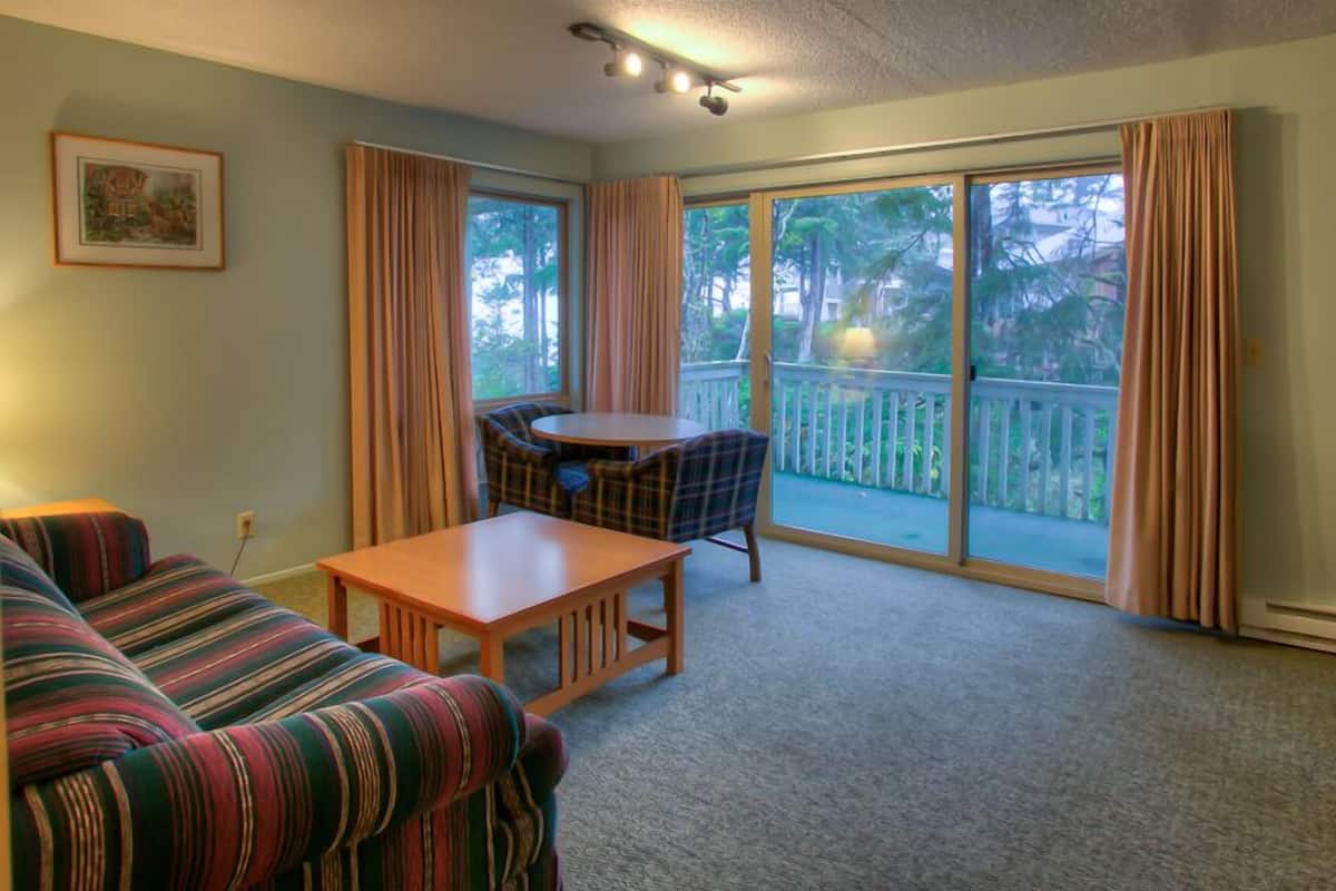 A living room with a sofa, a table, and a chair. There is a balcony with a view of trees and a white fence.