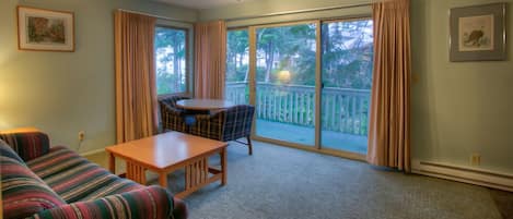 A living room with a sofa, a table, and a chair. There is a balcony with a view of trees and a white fence.