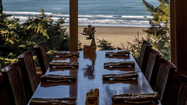Mesa de comedor de madera dispuesta para seis personas, con un florero en el centro, con vista a una playa con olas y árboles al fondo.