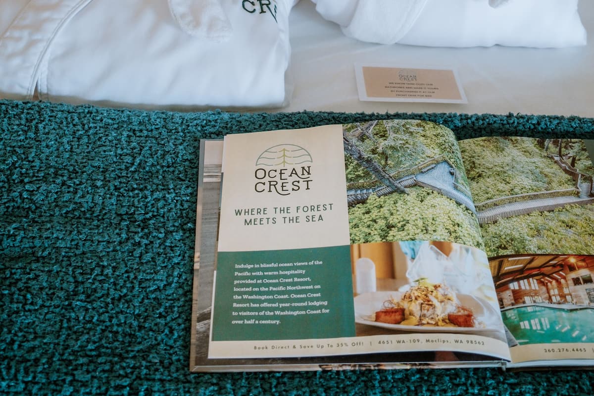 A neatly made bed with white bedding and two white towels tied with a bow, placed on a green textured area rug. A brochure titled "OCEAN CREST" is visible on the bed.