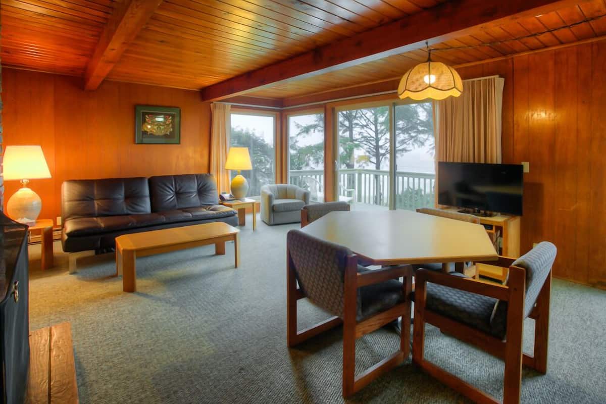 A cozy living room with a sofa, a coffee table, a dining table with chairs, a TV, and a large window with a view of trees.