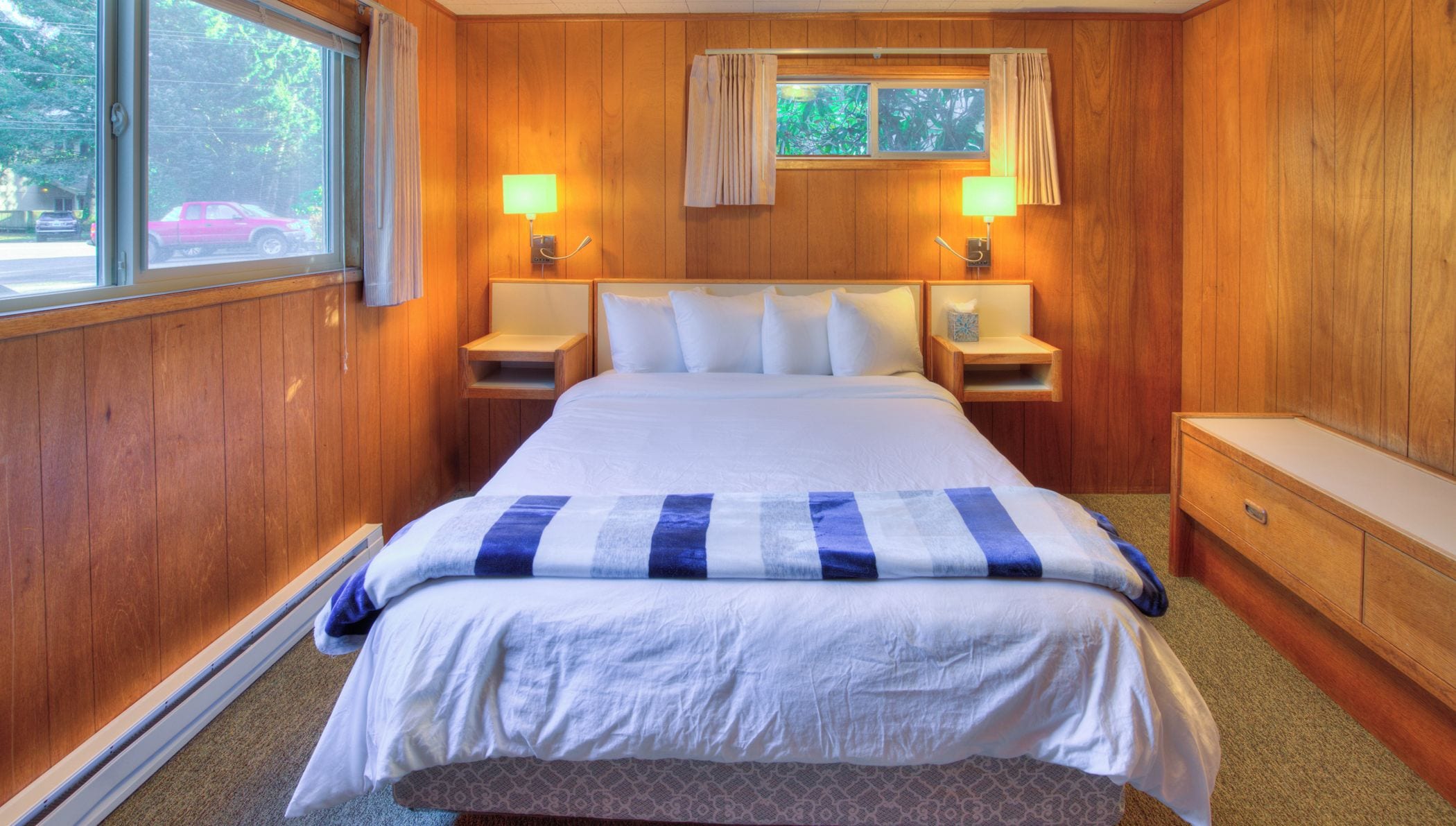 A hotel room with a large bed, two bedside tables, a nightstand, a window with curtains, and a small table with a lamp.