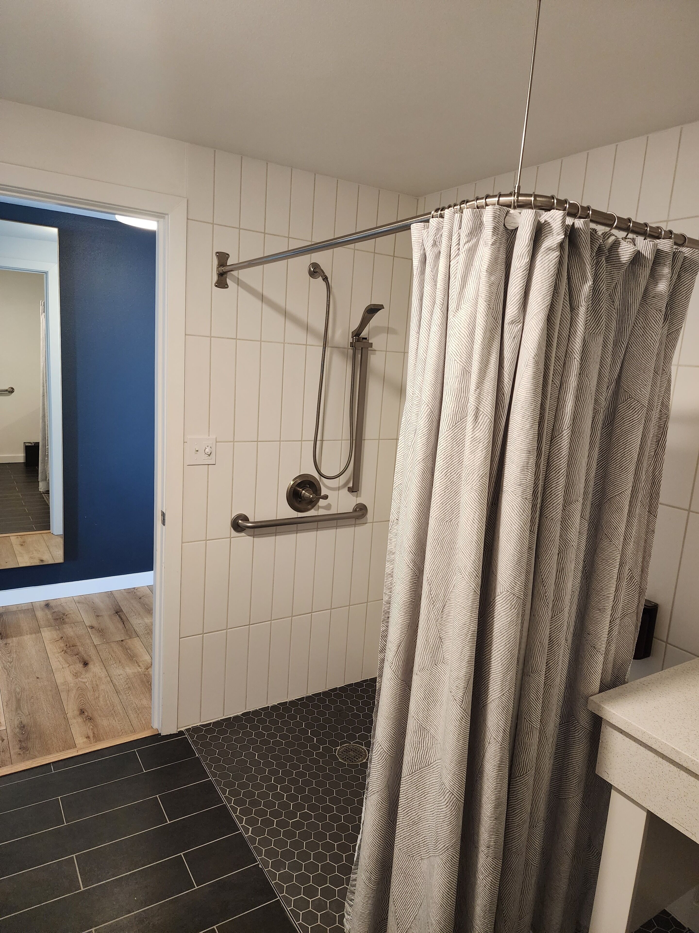 A shower with a patterned curtain, a handheld showerhead, and a handheld faucet. The shower is adjacent to a bathroom with a tiled floor, a sink, and a mirror.