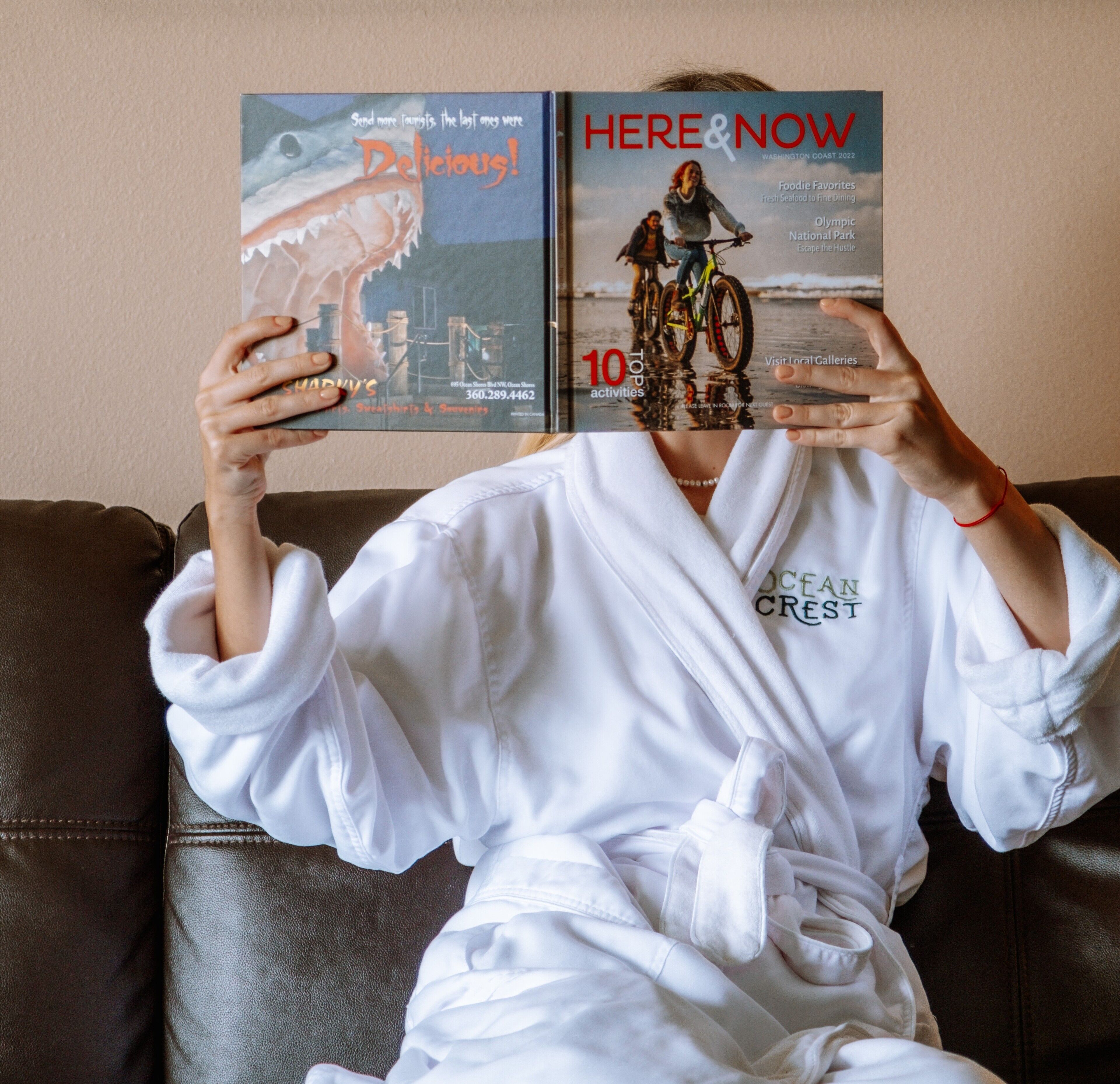 A person sitting on a brown leather couch, holding two books titled 'Delicious!' and 'Here & Now'. The person is wearing a white robe with 'OCEAN CRESCENT' embroidered on it.