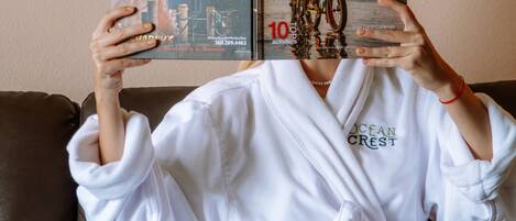A person sitting on a brown leather couch, holding two books titled 'Delicious!' and 'Here & Now'. The person is wearing a white robe with 'OCEAN CRESCENT' embroidered on it.