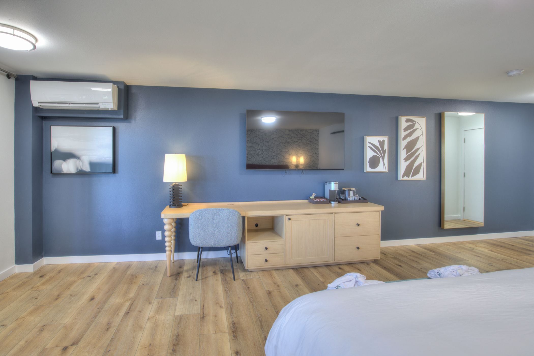 A hotel room with a bed, a desk with a chair, a wardrobe, a mirror, and artwork on the walls.