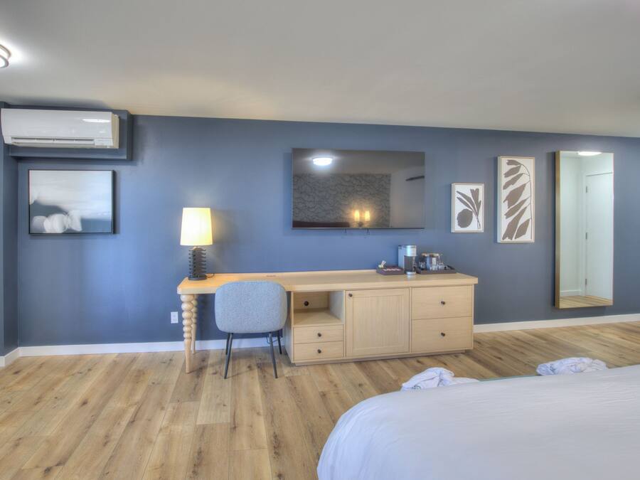 A hotel room with a bed, a desk with a chair, a wardrobe, a mirror, and artwork on the walls.