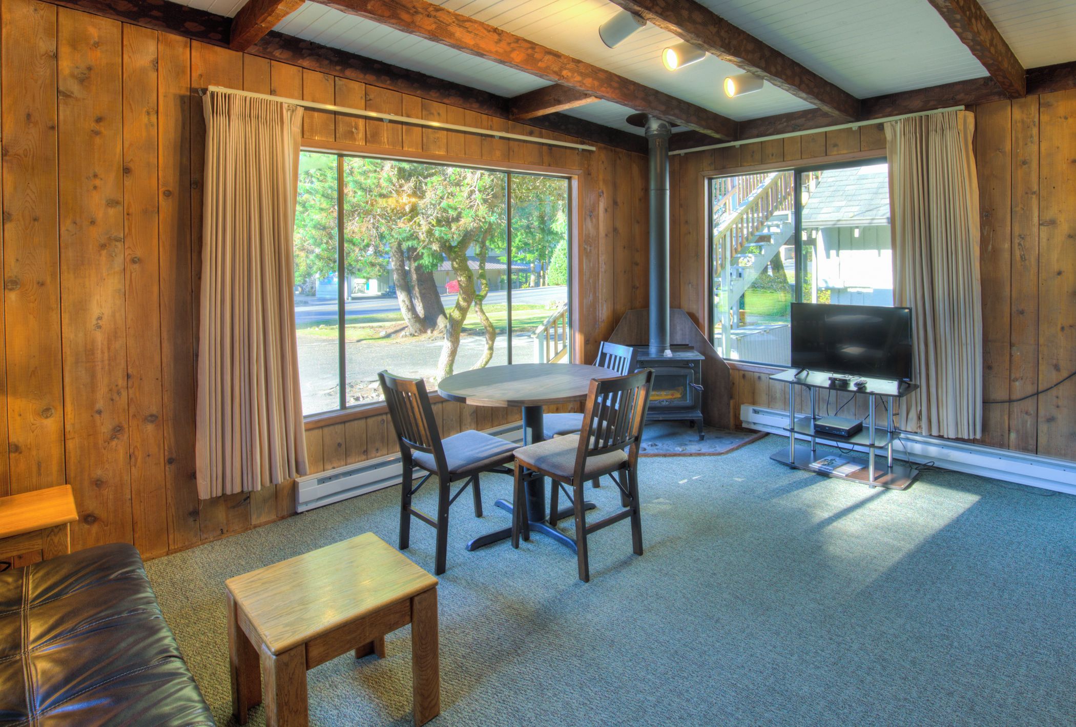 Una habitación con interiores de madera, una mesa con sillas, una chimenea, un televisor y ventanas grandes con cortinas.