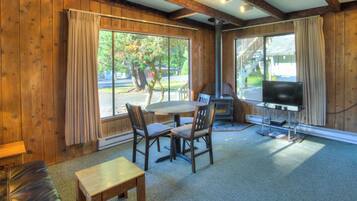 Una habitación con interiores de madera, una mesa con sillas, una chimenea, un televisor y ventanas grandes con cortinas.