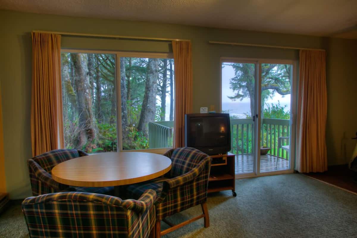 A living room with a round wooden table, plaid chairs, a television, and a sliding glass door leading to a deck with a view of trees and a body of water.