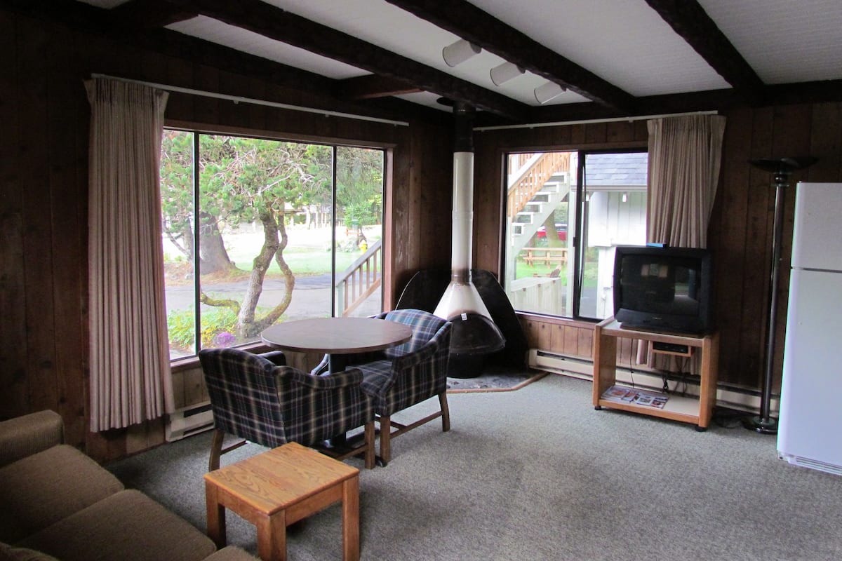 A cozy living room with a wooden ceiling, a round table, plaid chairs, a sofa, a television on a stand, and a refrigerator.