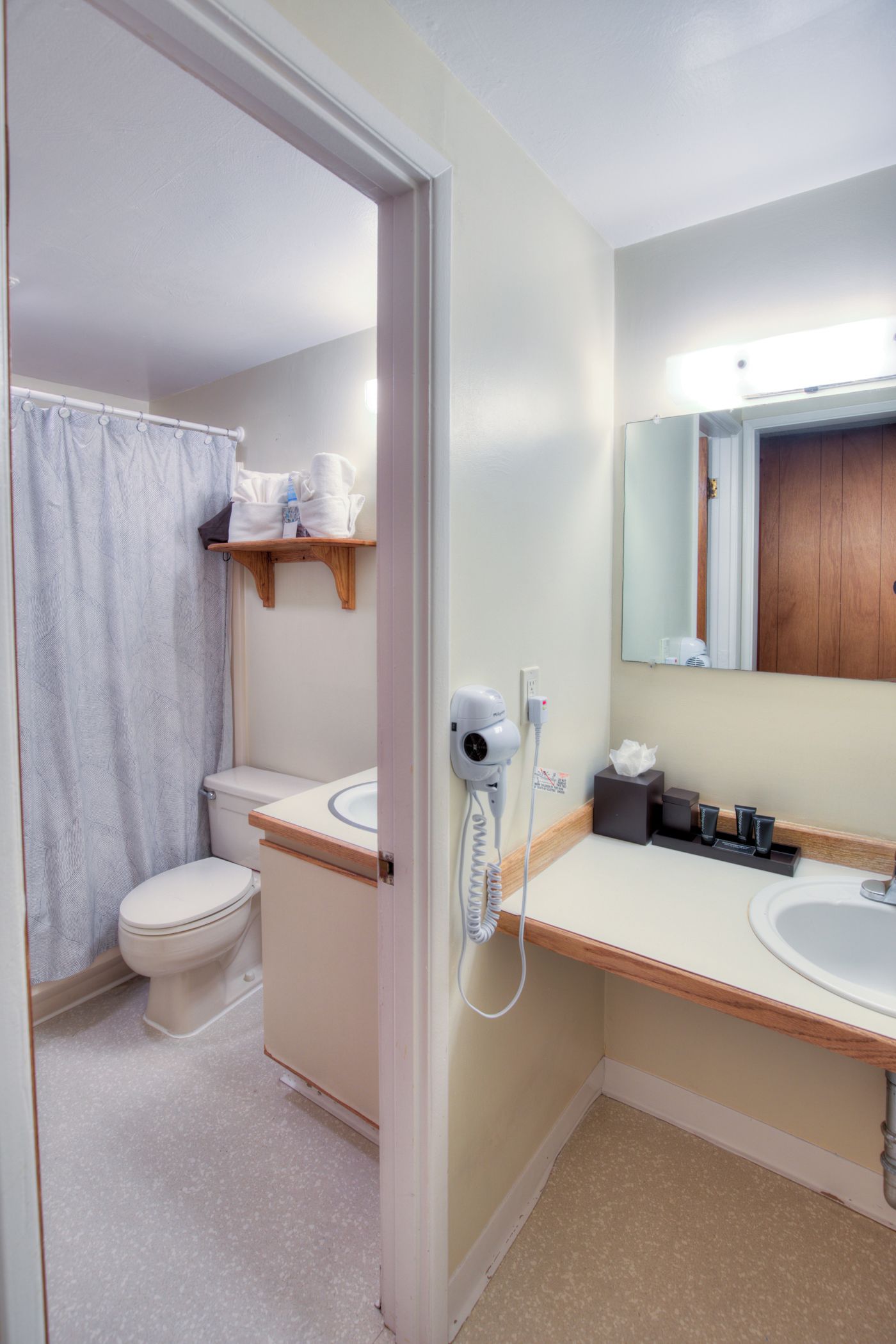 Un baño con inodoro, cortina de ducha, lavabo con espejo y secador de pelo en la pared.