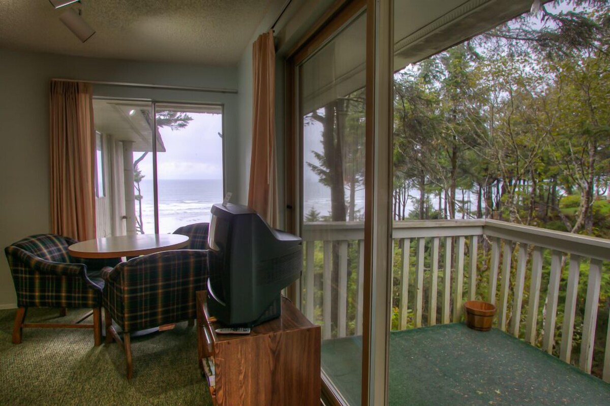Un balcón con vista al océano, una mesa con sillas y un televisor sobre un atril de madera.