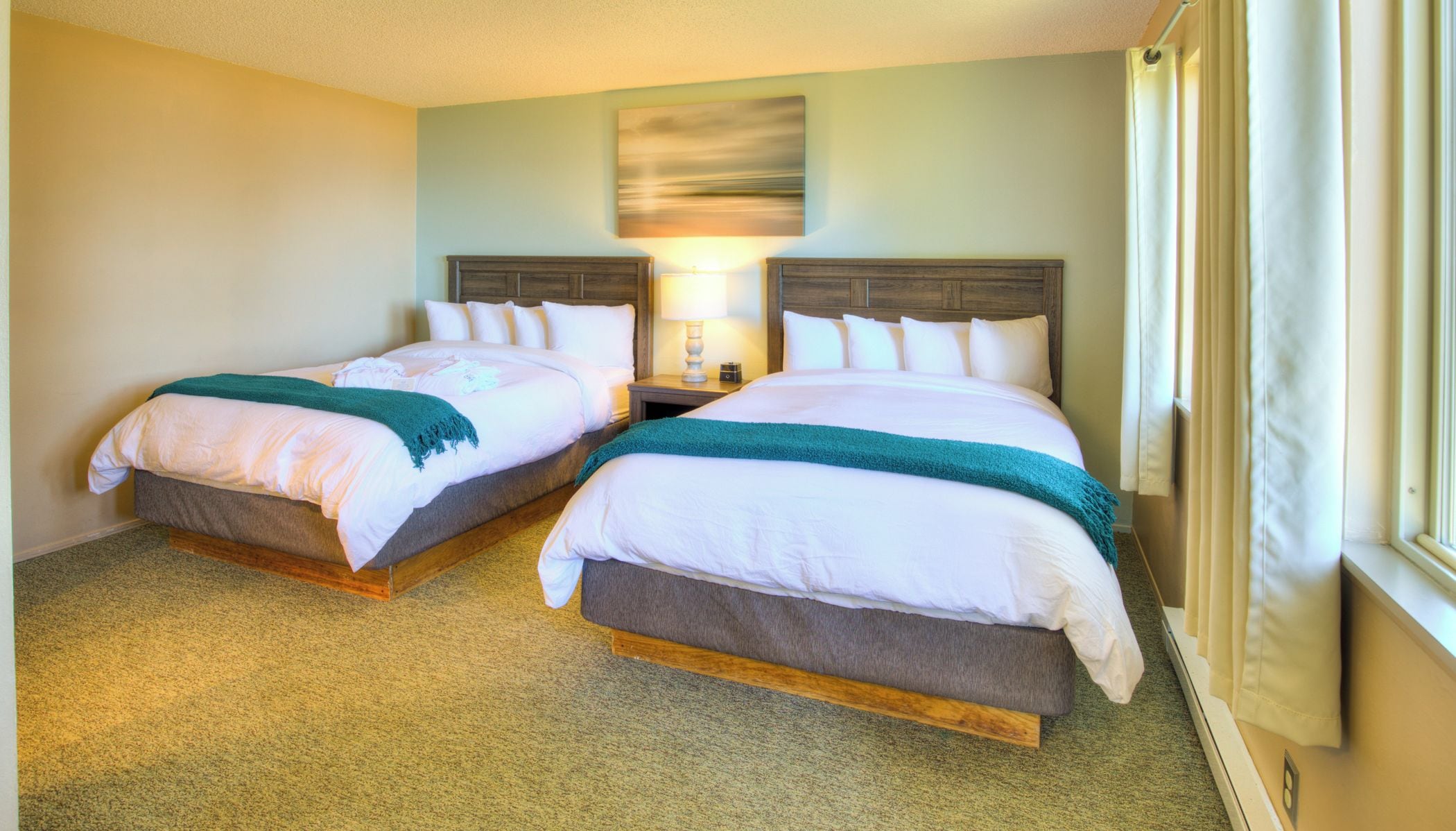 A hotel room with two beds, a nightstand with a lamp, a painting on the wall, and a window with curtains.