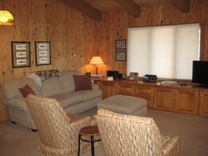 A cozy living room with a wooden interior, a sofa, two patterned armchairs, a coffee table, a television, and framed pictures on the wall.
