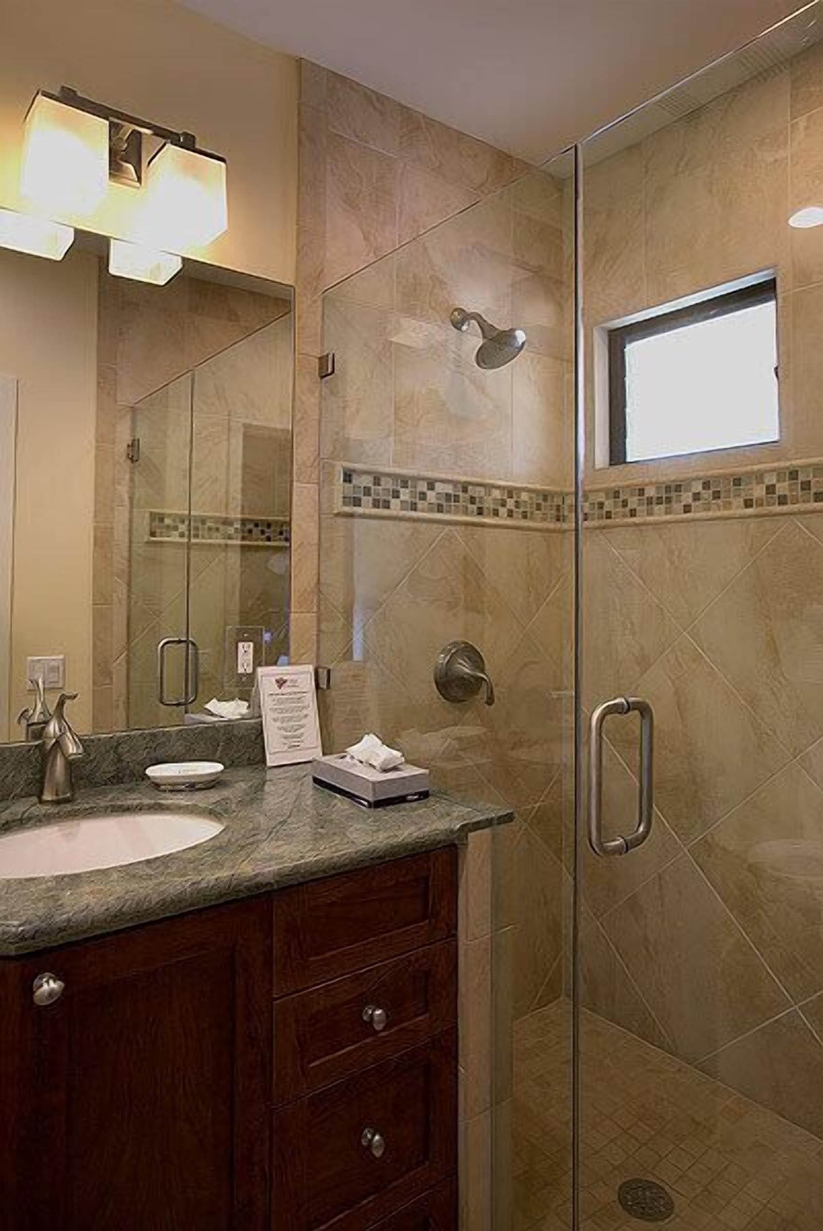 A bathroom with a glass-enclosed shower, a sink with a marble countertop, and a mirror with a light fixture.