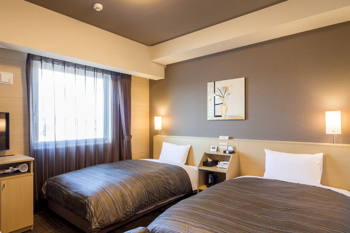 A hotel room with two beds, a desk with a lamp, a TV, and a window with curtains.