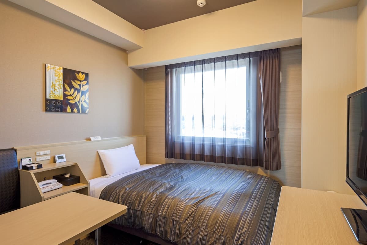 A hotel room with a bed, a desk, a television, a window with curtains, and a painting on the wall.