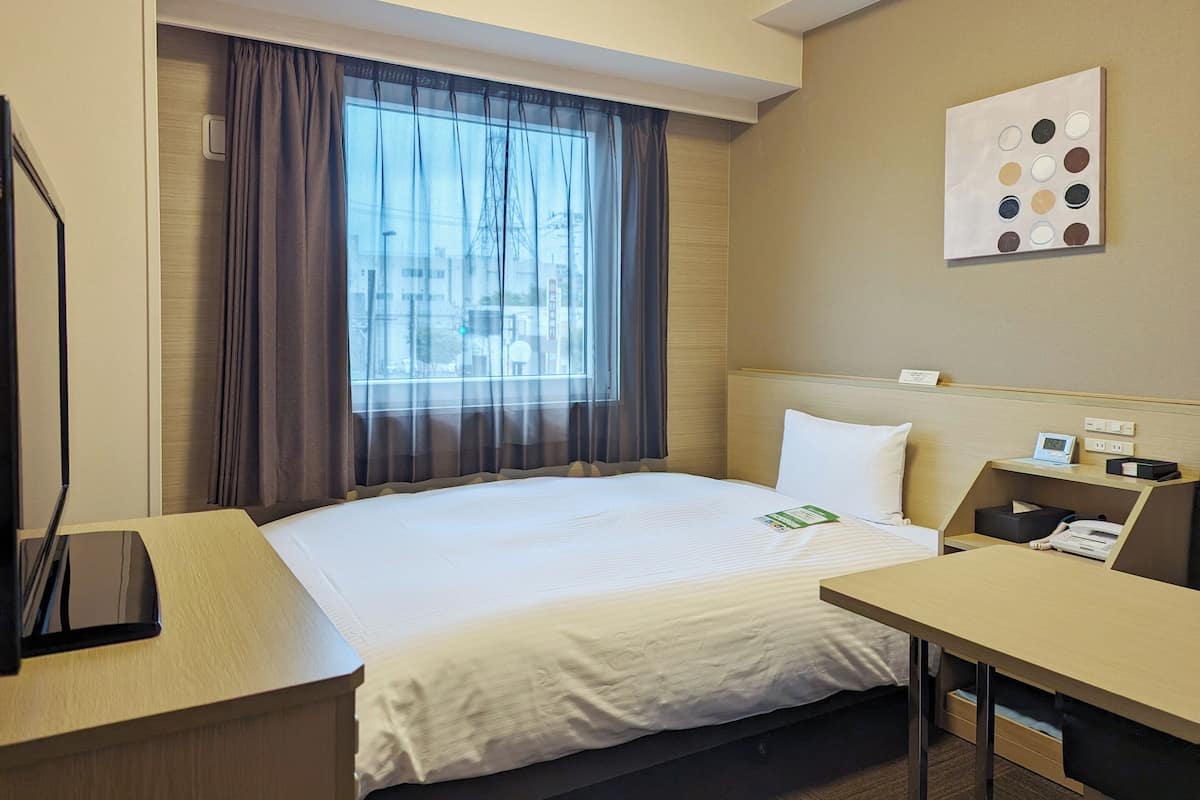 A hotel room with a bed, a desk, a chair, a window with curtains, a wall clock, and a painting.