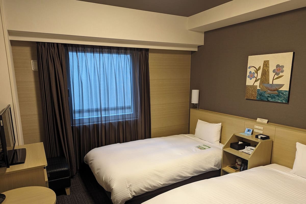 A hotel room with two beds, a desk, a chair, a small table, a window with curtains, and a wall-mounted artwork.