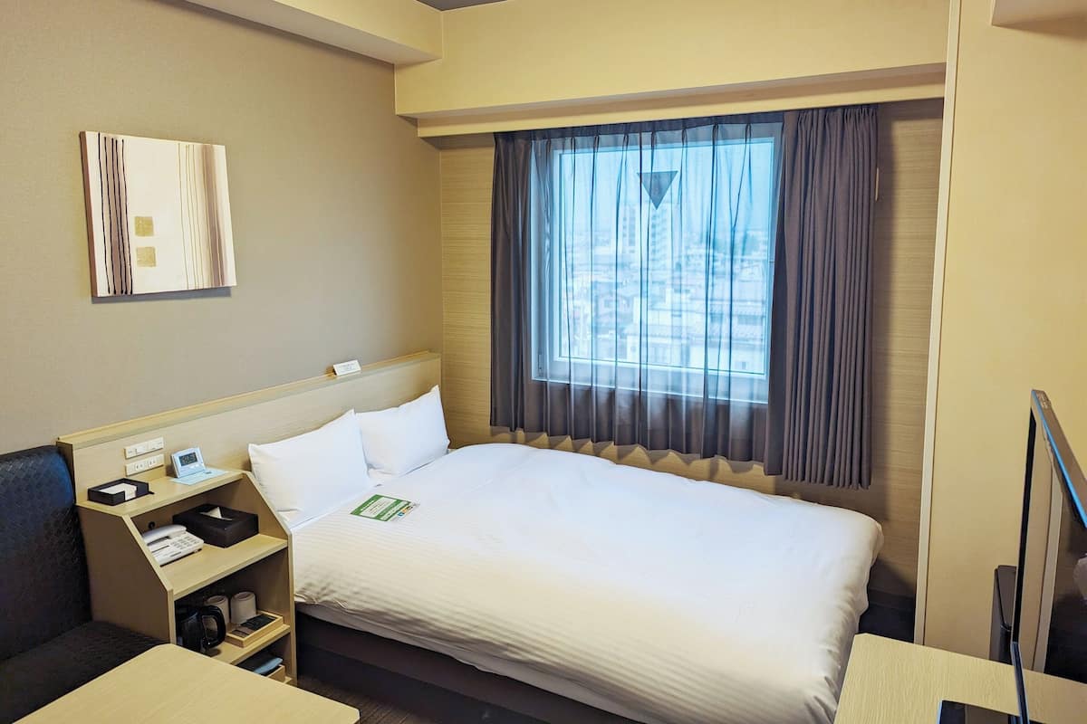 A hotel room with a bed, bedside table, and a window with curtains.