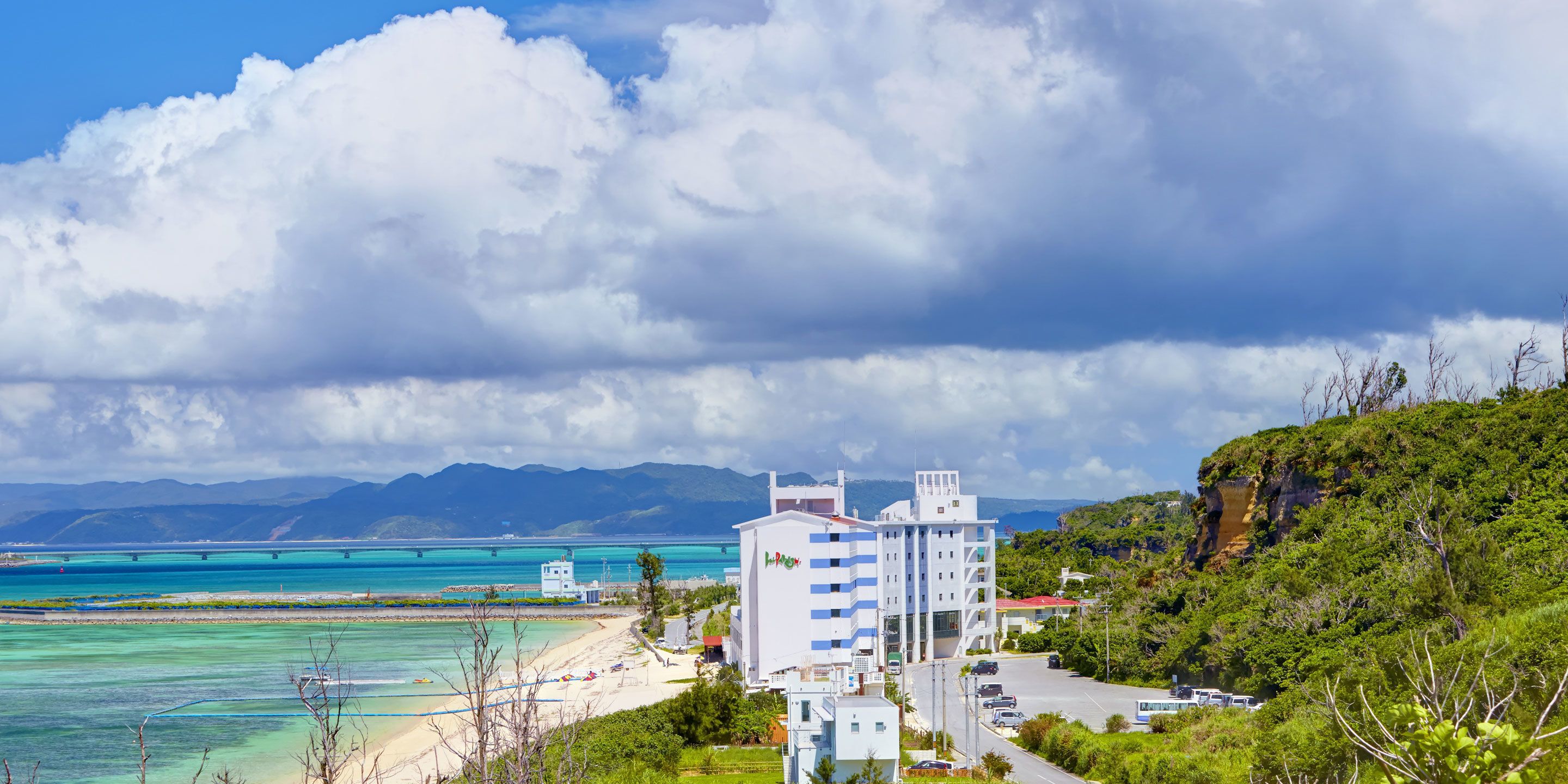 A coastal hotel with a beach view, a parking area, and a mountainous backdrop.