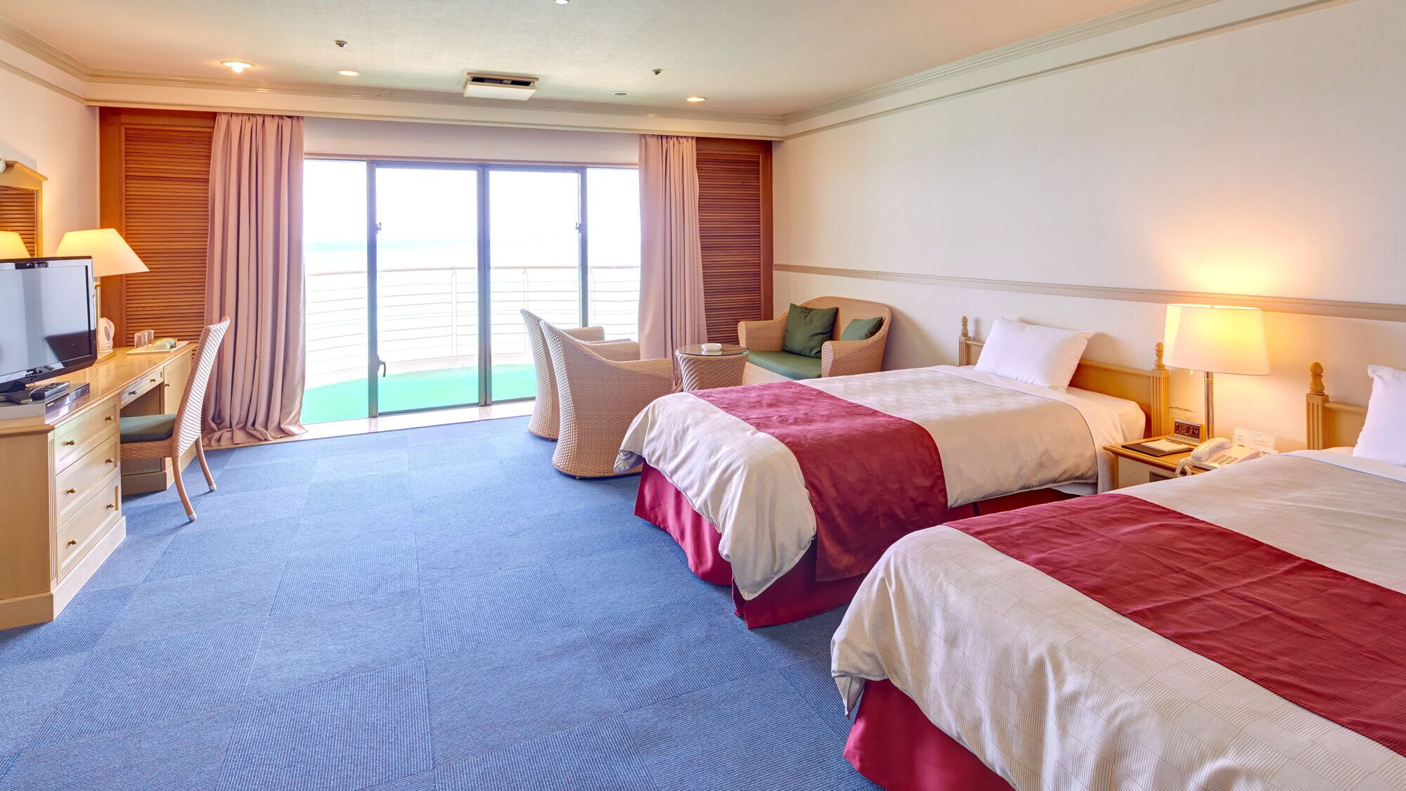 Ein Hotelzimmer mit zwei Betten, einem Schreibtisch, einem Stuhl, einem kleinen Tisch und Blick ins Freie.