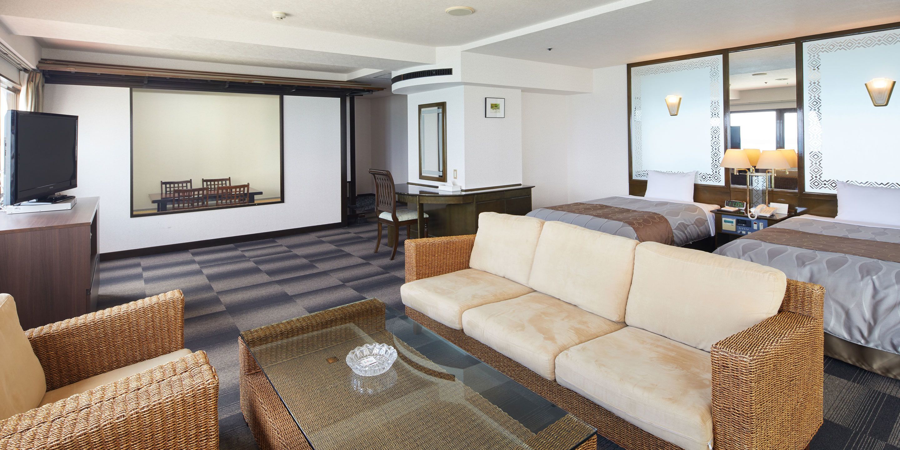 A hotel room with a sofa, a glass coffee table, a TV, a desk, and a bed.