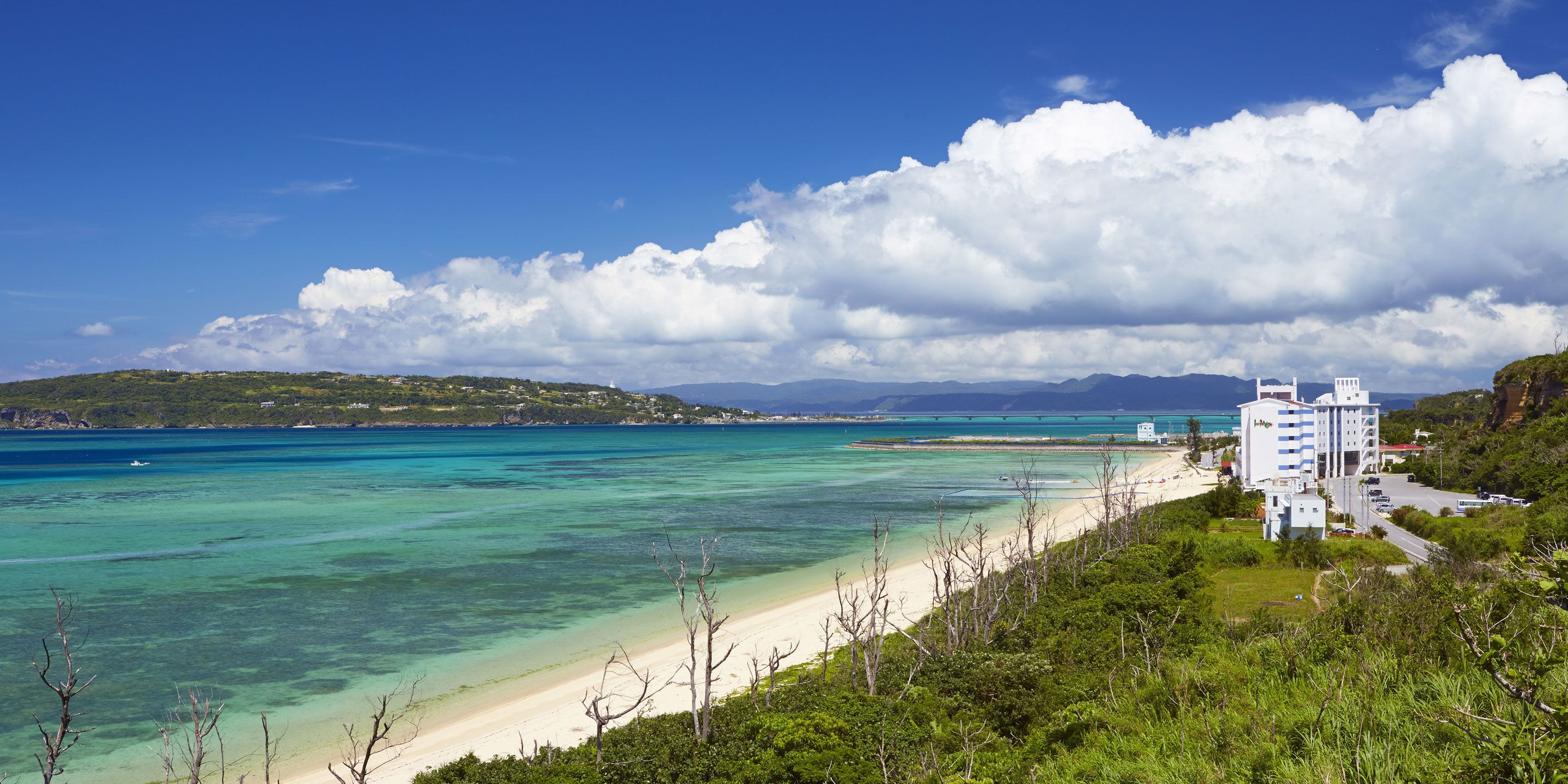 A coastal hotel with a beach view, a clear sky, and a mountainous backdrop.
