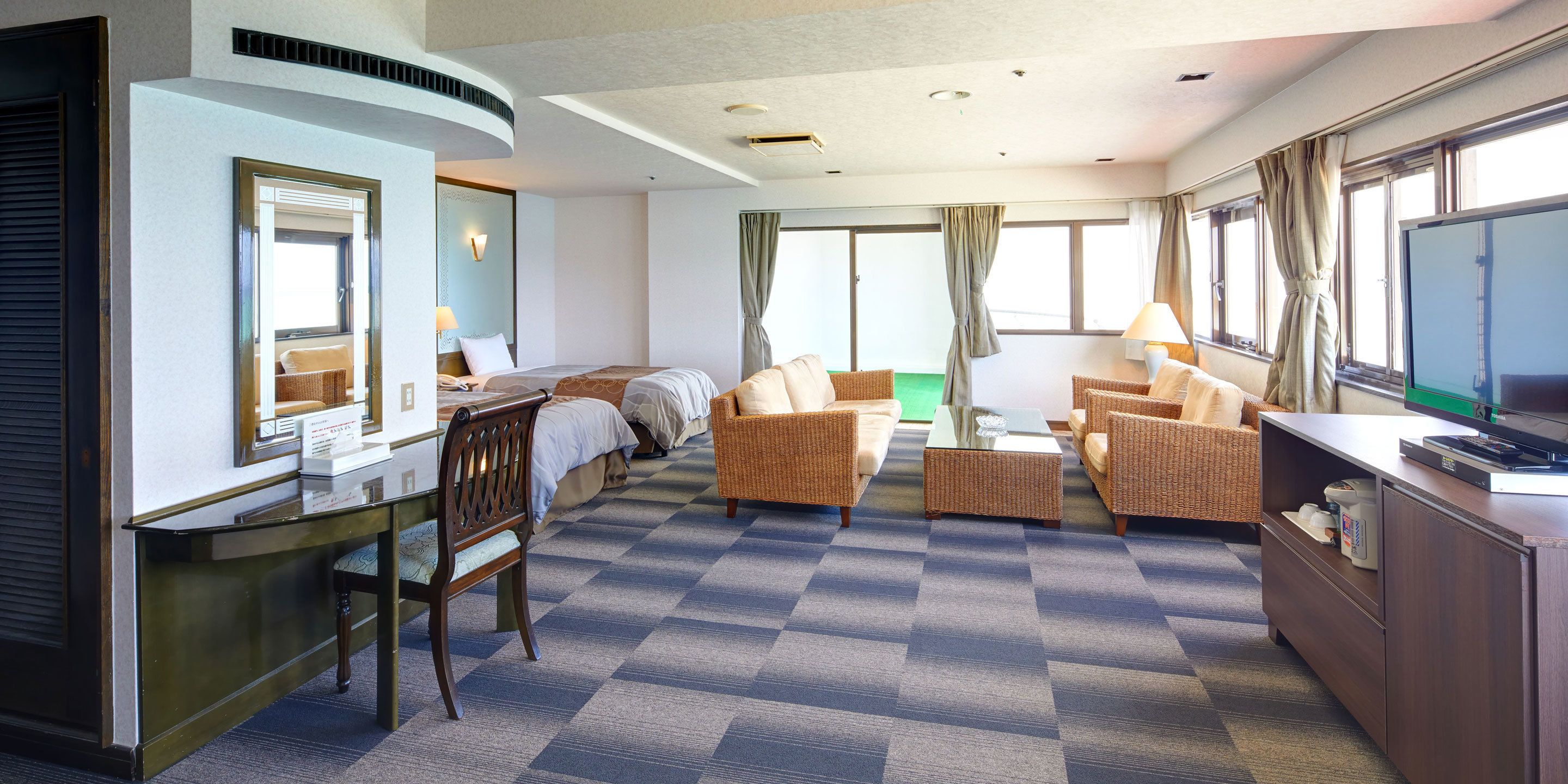 A hotel room with a bed, a desk, a television, wicker furniture, and large windows.
