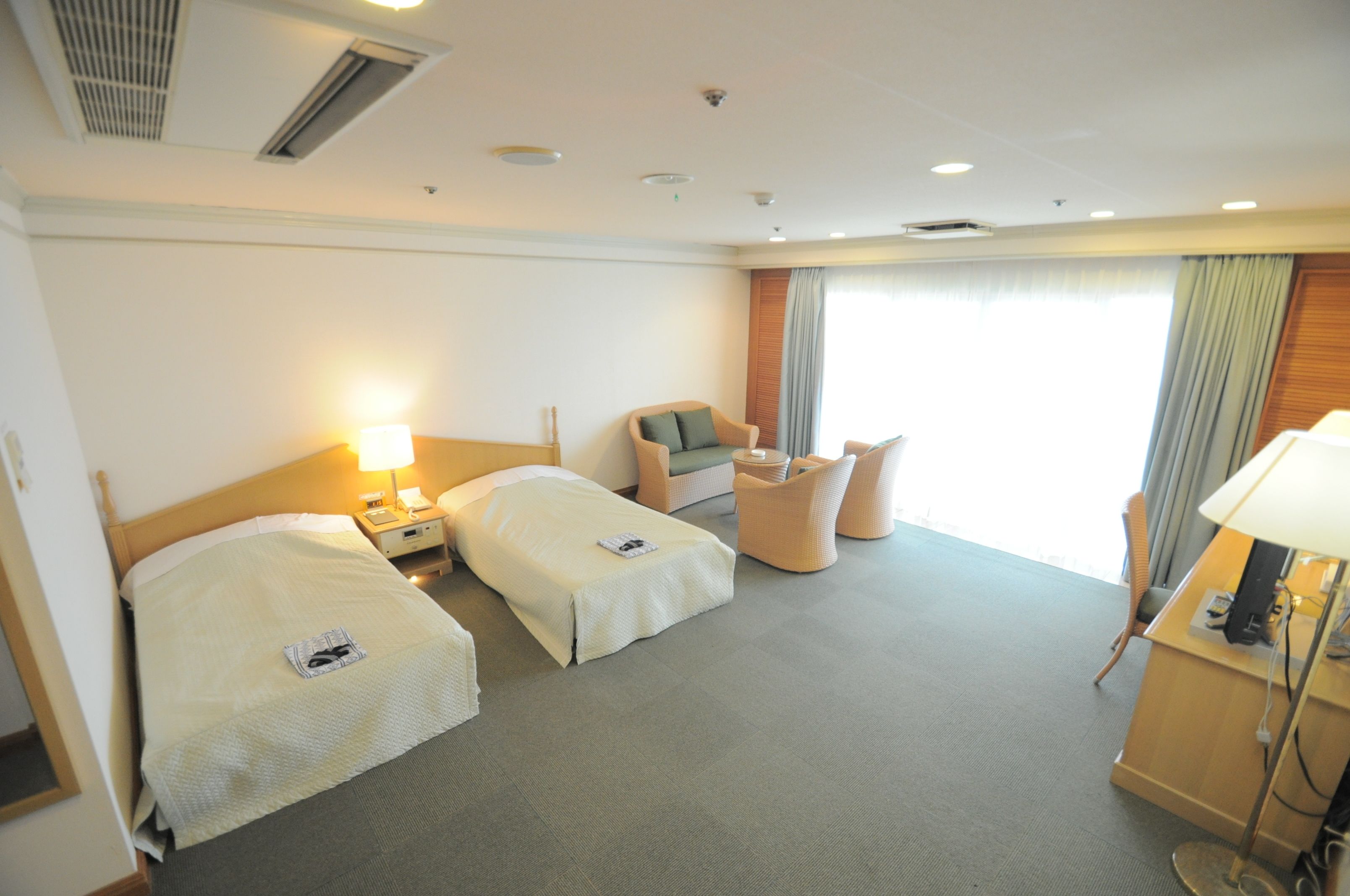 Habitación de hotel con dos camas individuales, un pequeño sofá, un escritorio con lámpara y un gran ventanal con cortinas.
