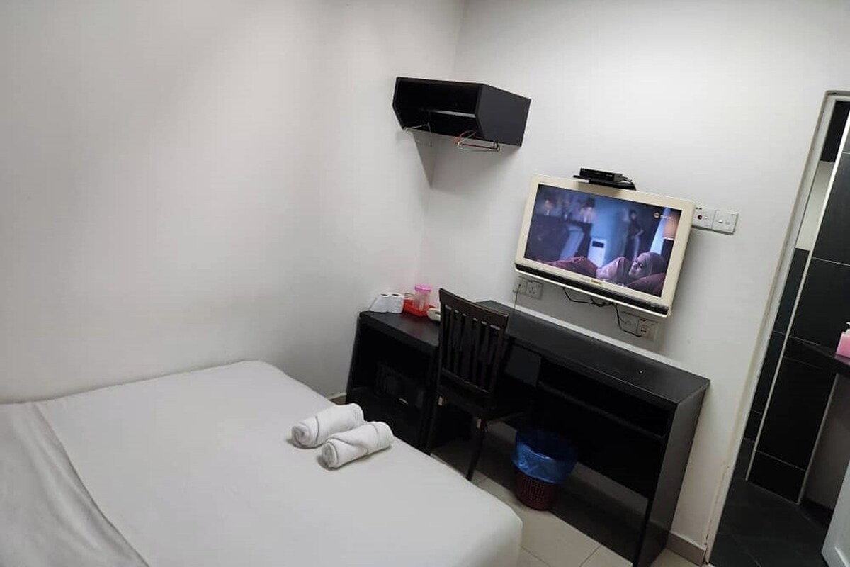 A hotel room with a bed, a desk with a television, a chair, and a small trash bin.