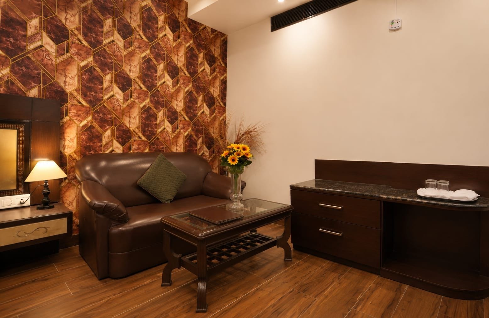 A hotel room with a leather sofa, a small coffee table, a vase with flowers, a minibar, and a wall with a geometric pattern.