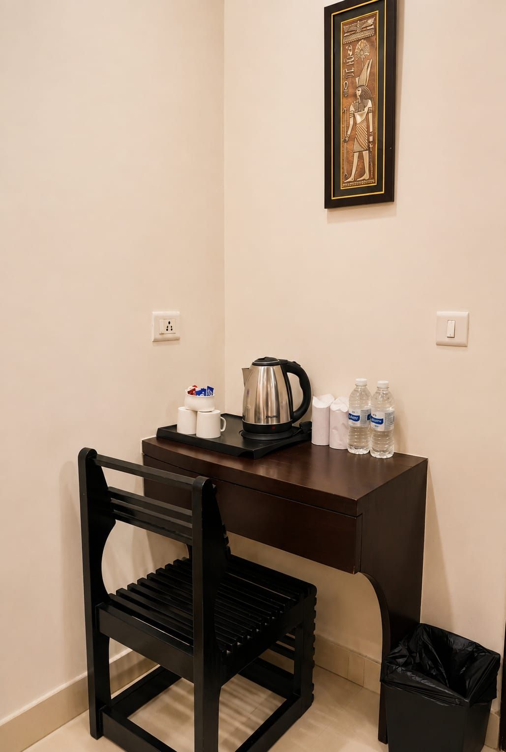 A small room with a dark wooden desk, a black chair, a framed picture on the wall, a kettle, cups, and bottles.