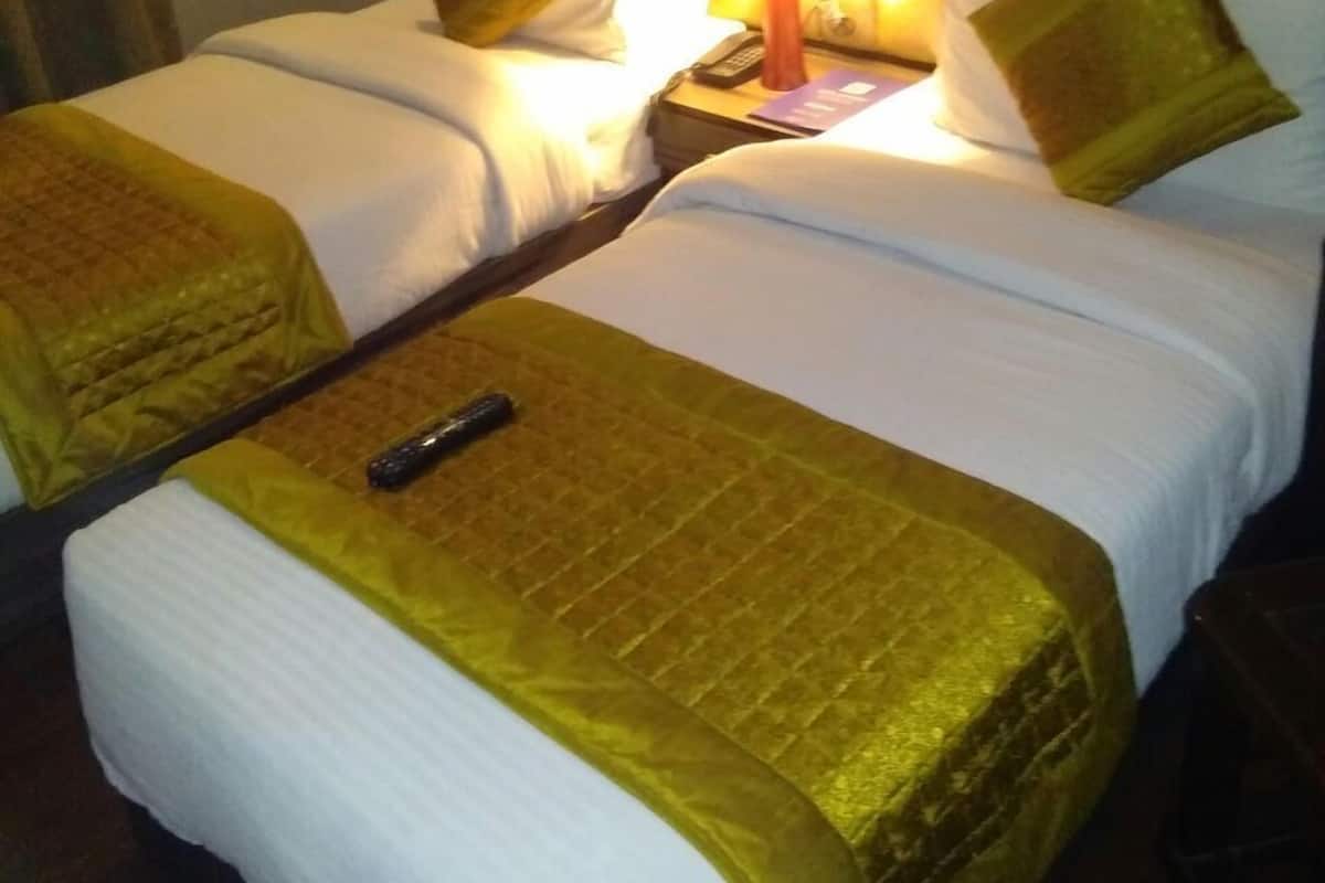 A hotel room with two beds, each with white linens and a gold throw blanket. There is a bedside table with a lamp, a remote control, and a book.