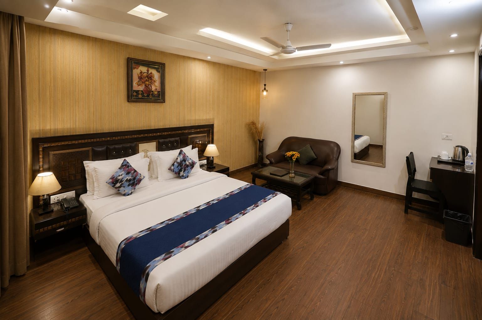 A hotel room with a large bed, bedside tables, a desk, a chair, a mirror, and a ceiling fan.