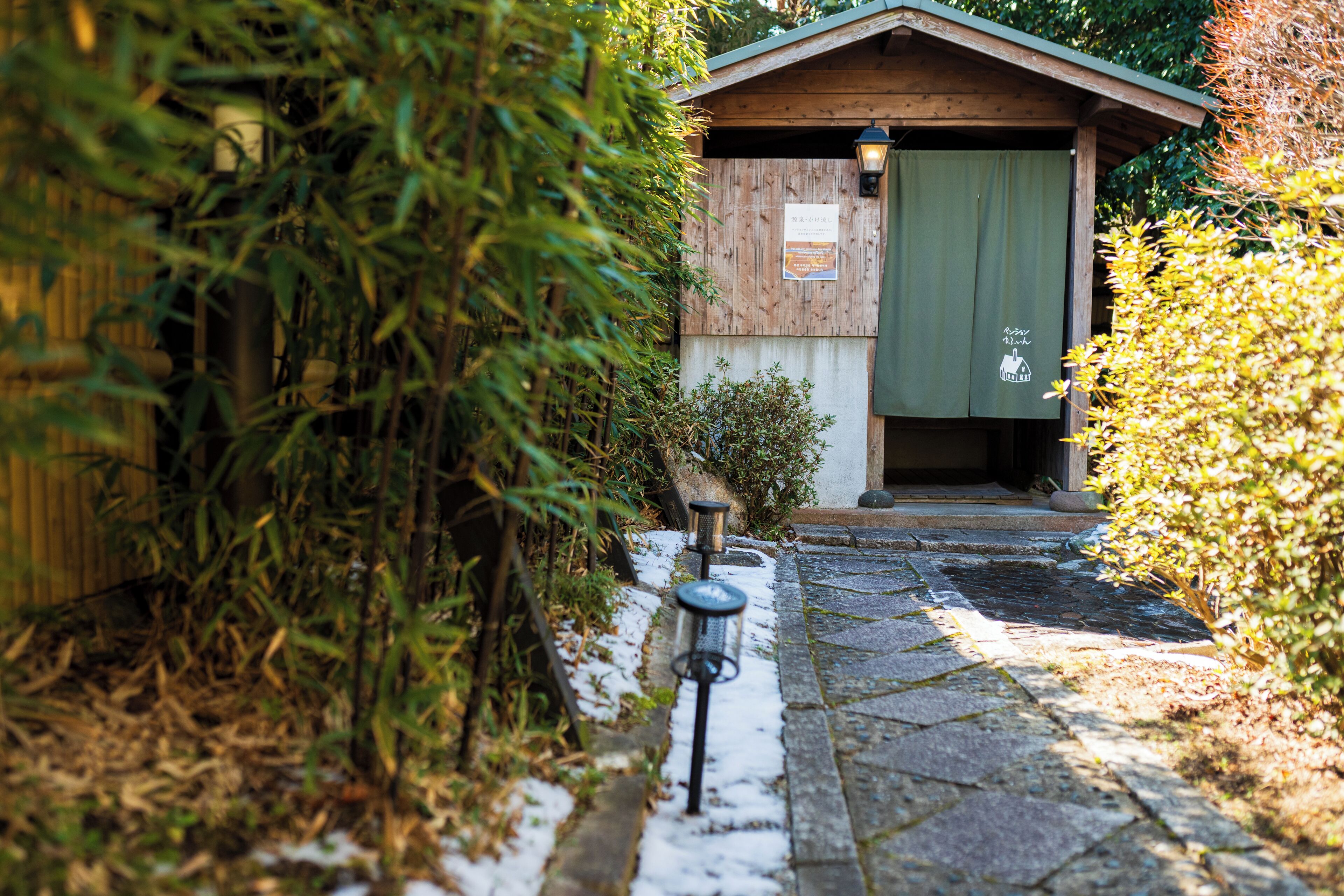 A stone pathway leading to a wooden building with a green door, surrounded by greenery and a snow-covered ground.