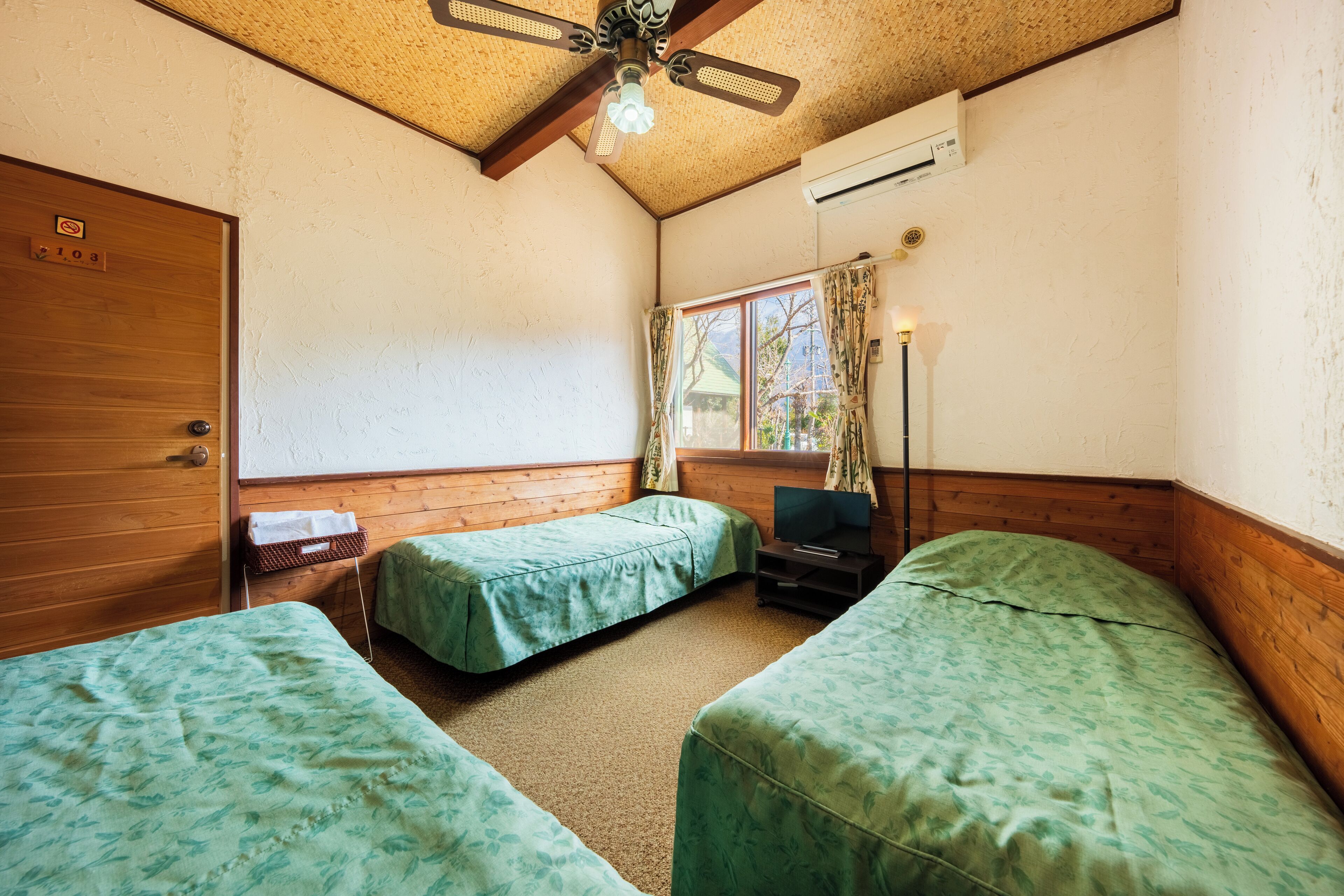 A room with two beds, a wooden door, a window with curtains, a ceiling fan, and an air conditioning unit.
