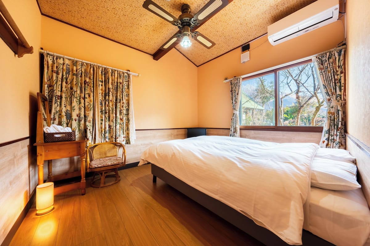Un dormitorio con cama, una silla, mesita de noche, ventilador de techo y ventana con cortinas.