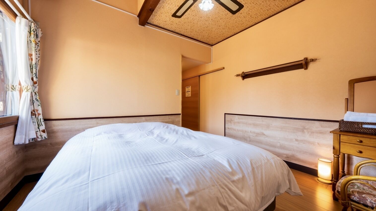 Un dormitorio con una cama, cabecera de madera, una mesita de noche, una lámpara y una ventana con cortinas.