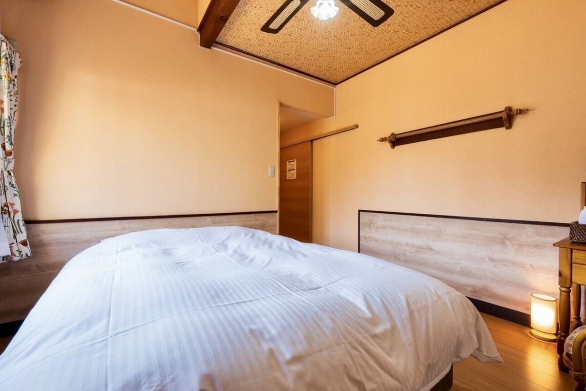 Un dormitorio con una cama, cabecera de madera, una mesita de noche, una lámpara y una ventana con cortinas.