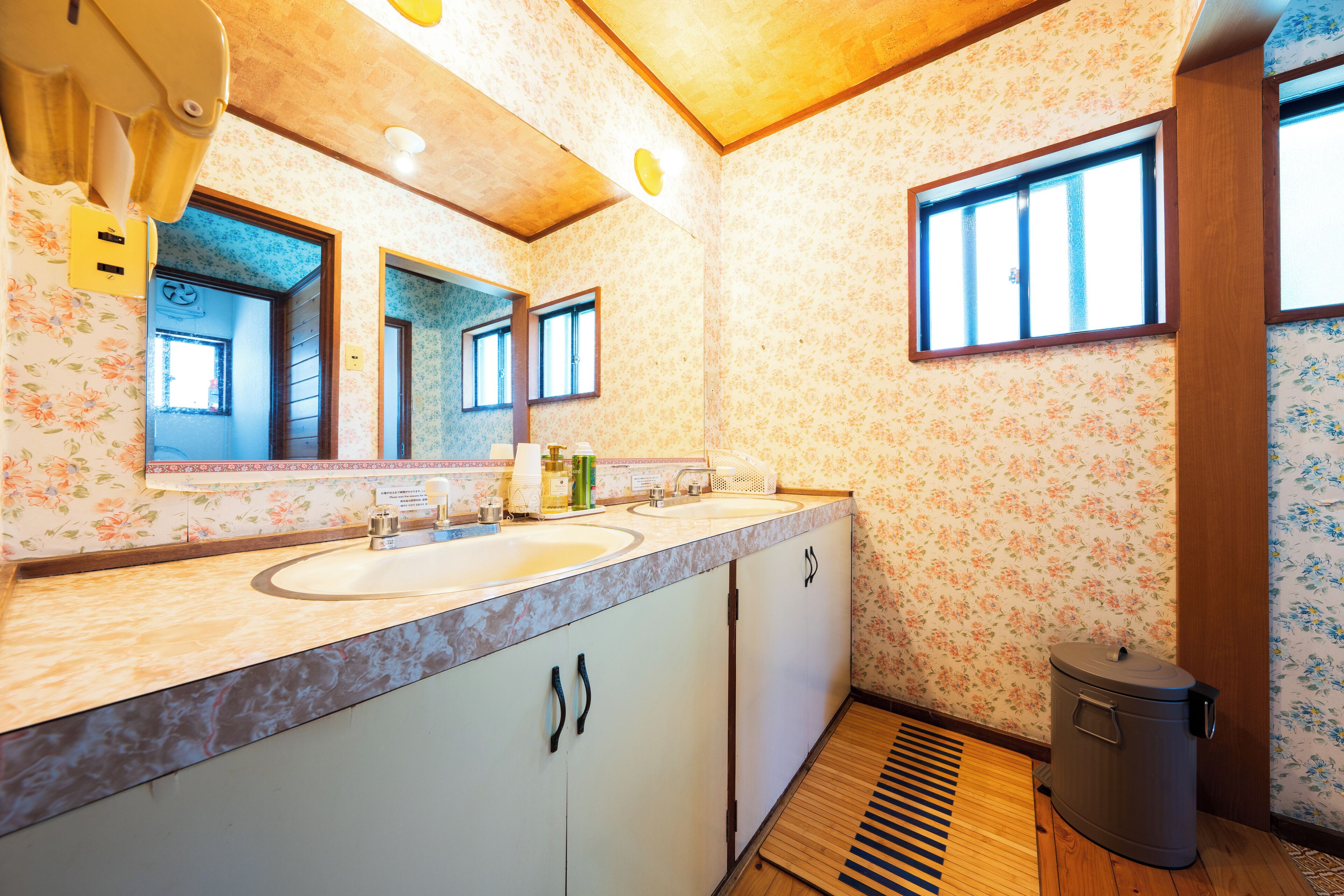 Un baño con lavamanos doble, piso de madera y un bote de basura.