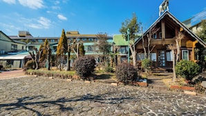 A stone-paved courtyard with a wooden building featuring a statue on the roof, surrounded by trees and shrubs. - Pension Yufuin (Yufu)