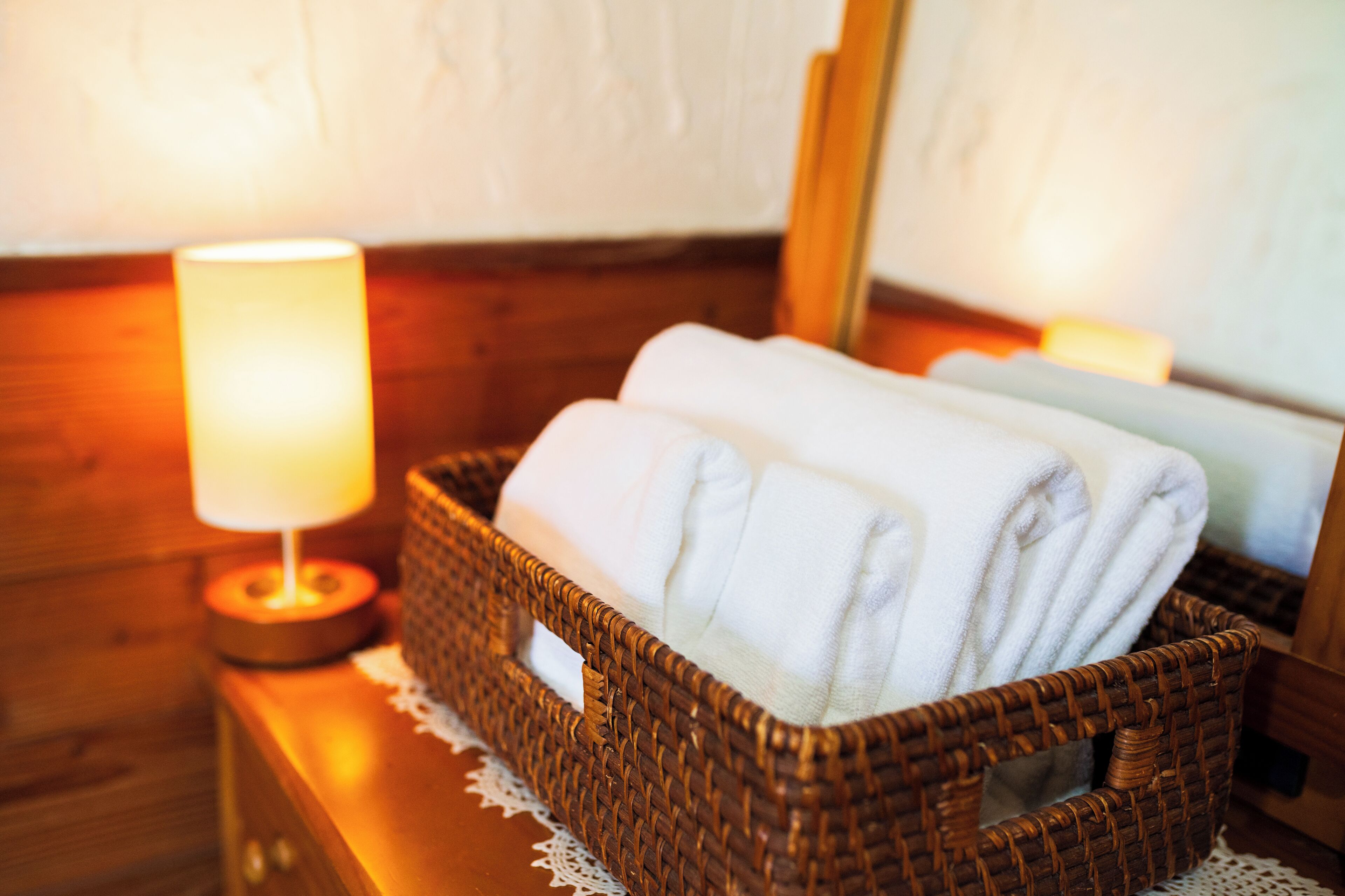 A wicker basket with folded towels on a wooden nightstand next to a lit lamp.