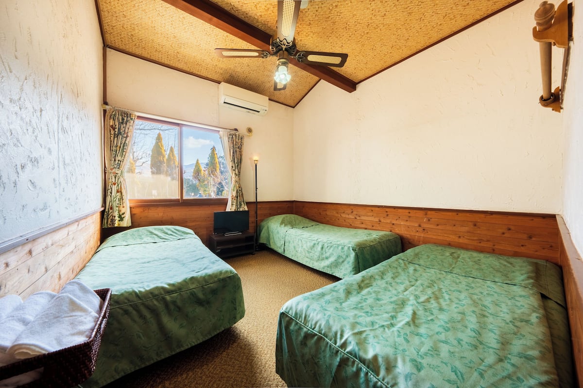 Una habitación con dos camas individuales, un ventilador de techo, una ventana con cortinas y una mesita de noche con lámpara.