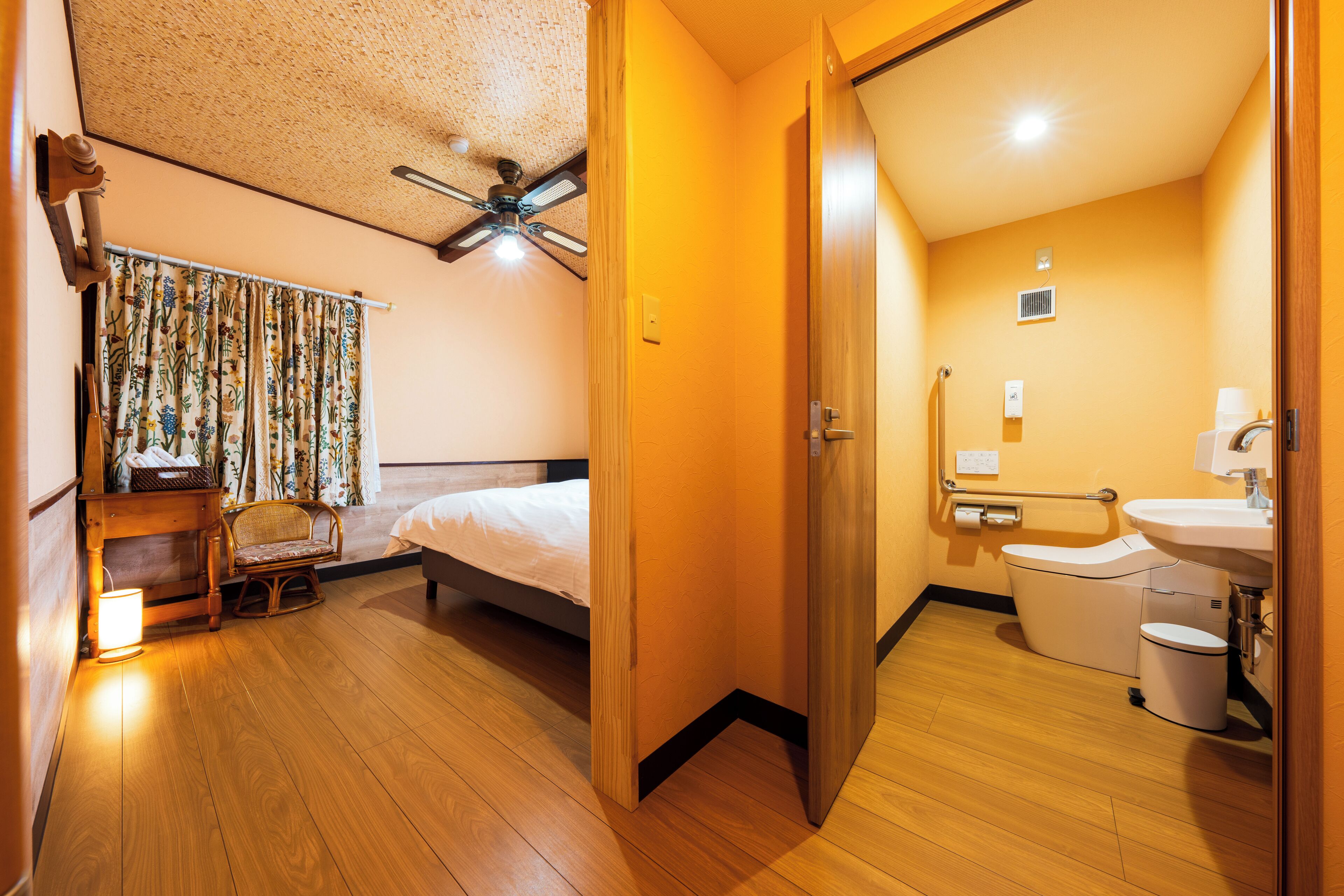 Una habitación de hotel con cama, ventilador de techo, baño con inodoro y lavamanos, y piso de madera.