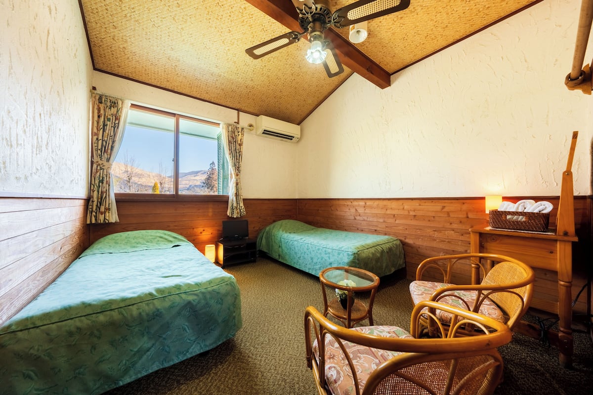 Una habitación con dos camas individuales, techo de madera, ventana con cortinas, una mesita con una silla y un calentador fijado a la pared.