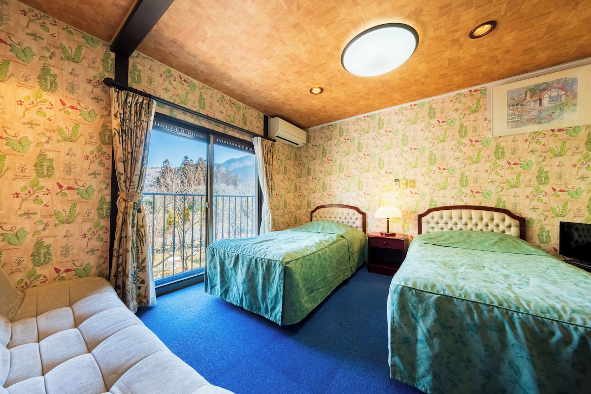 Una habitación con dos camas, un sofá, una ventana con cortinas, una lámpara en el techo y un papel tapiz con motivos botánicos.