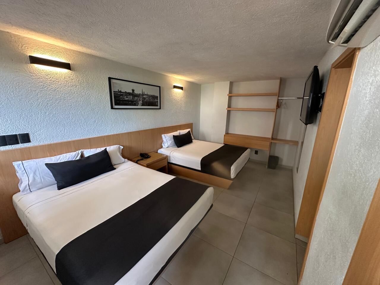 A hotel room with two beds, a flat-screen TV, a small table, and a wall-mounted shelf.