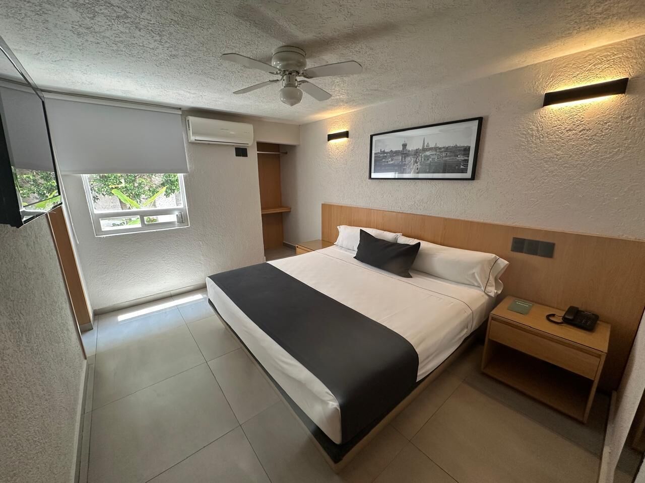 A hotel room with a bed, a nightstand, a ceiling fan, a framed picture on the wall, and a window.